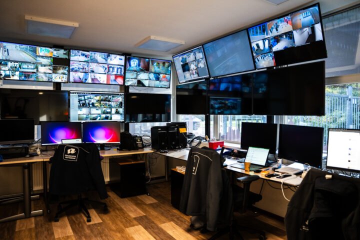 Foto van de beveiligingkantoor van de hogeschool. Een klein ruimte met 12 grote monitors die aan het plafon hangen waarop camera beelden van alle locaties streamen. En bureau werkplekken voor 3 medewerkers.