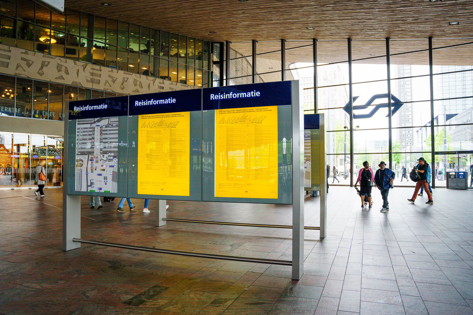 Foto van gele NS-reisinformatiezuilen op Rotterdam Centraal. Op de achtergrond het NS-logo op de glazen wand van de buitengevel.