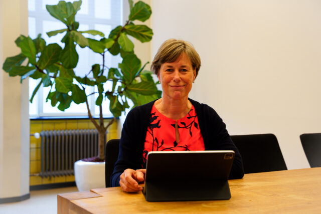 Foto van Hanneke in haar kantoor aan tafel achter haar Surface computer. Hanneke draag een rode jurk met zwarte patroon van planten en een zwarte cardigan