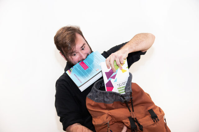 portretfoto van Jonnick voor een witte muur. Met paniekerige uitdrukking probeert hij boeken in een rugzak te proppen, een van de boeken tussen zijn tanden geklemd.