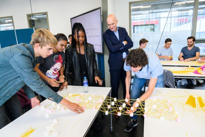 Minister Kuipers kijkt aandachtig naar de door studenten gemaakte brug van spaghetti en marsmallows