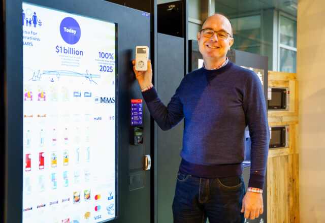 Foto van Wouter Sjoerdsma naast snackautomaat met een grote display. Wouter houdt een elektriciteitsmeter vast.