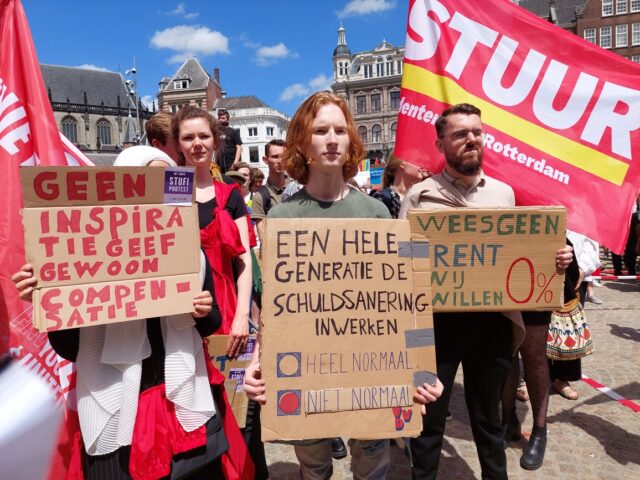 Foto van protesterende studenten met STUUR vlaggen en protestborden in Amsterdam. De borden lezen onder anderen "Geen inspiratie geef gewoon compensatie" en "Wees geen krent, wij willen nul procent".